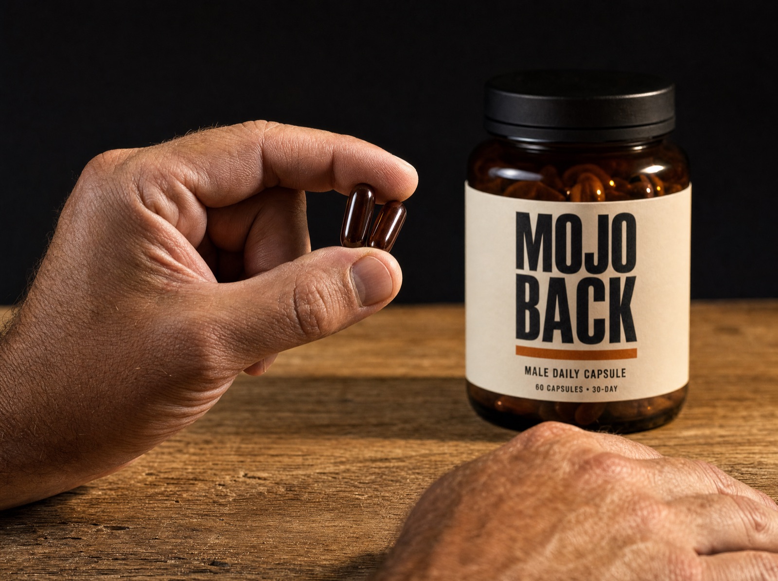 A man's hand holding two capsules beside the MOJO BACK jar on a wooden counter, morning light